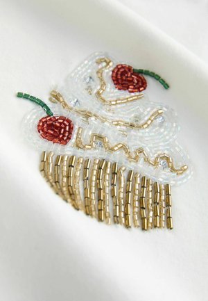 Beaded cupcake embroidery with red cherries, green stems, and cascading gold beads on a white fabric background. Textured and colorful design.