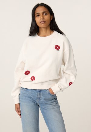 Vrouw met lang donker haar draagt een witte sweatshirt met rode lippenafdrukken en lichtblauwe spijkerbroek, staat met één hand in de zak.