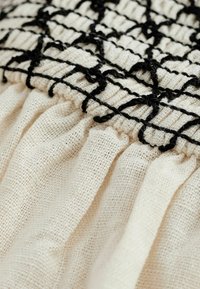 Gros plan d'un tissu beige tissé avec un motif en grille brodé en noir et un détail de couture froncée.