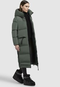 Cappotto imbottito lungo di color verde scuro con colletto alto, imbottitura verticale e zip frontale. Abbinato a stivali neri isolanti e pantaloni aderenti.