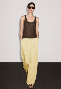 Bruine gebreide tanktop met brede schouderbanden, gecombineerd met gele linnen broek met wijde pijpen. Het model draagt zwarte zonnebril en bruine sandaal.