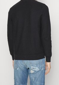 Svart stickad sweatshirt med rund halsringning, ribbade muddar och fåll. Bärs över ljusblå slitna jeans, visande bakifrån med texturerat tyg.