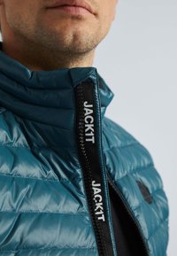 Veste matelassée teal avec un col montant, un motif matelassé horizontal, et une fermeture éclair noire avec le logo "JACK1T".