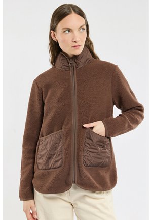 Femme portant une veste polaire marron zippée avec des poches matelassées et un col, regardant légèrement sur le côté, main dans la poche.