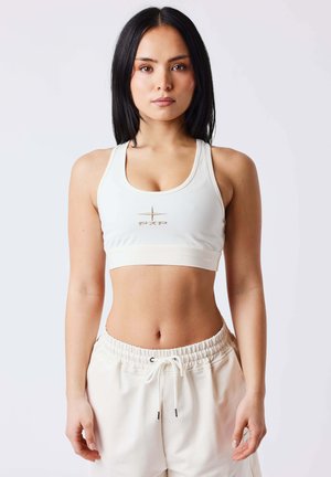 Femme aux longs cheveux noirs portant un brassière de sport blanche avec un logo et un pantalon blanc taille haute à cordon, debout devant un fond uni.