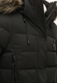 Chaqueta de invierno negra acolchada con capucha adornada con piel, ajustadores de cordón, botones de presión y bolsillo lateral con cremallera, llevada sobre un suéter blanco.
