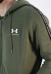 Sudadera verde hecha de un tejido suave con cremallera y capucha con cordón. Presenta un logo blanco en el pecho y rayas de acento negras en las mangas.