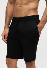 Torse masculin sportif portant un short noir à cordon avec les mains dans les poches, sur un fond uni gris clair.