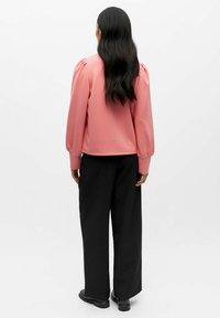 Roze blouse met lange mouwen en pompon-schouders, gecombineerd met zwarte wijde broek en zwarte schoenen. Eenvoudige, gladde textuur met een ontspannen pasvorm.