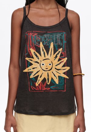 Mujer con una camiseta sin mangas negra con un gráfico de sol amarillo y diseños de fondo en rojo y verde azulado, combinada con pantalones beige claro.