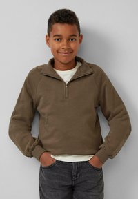 Bruine, lange mouwen pullover met een half-rits kraag en ribbenmanchetten, gedragen over een lichtgekleurde t-shirt, gecombineerd met donkergrijze jeans.