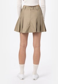 Jupe plissée kaki avec une taille cintrée, fermeture éclair et bouton. Tissu léger avec un ourlet fluide. Portée avec des baskets blanches et des chaussettes.