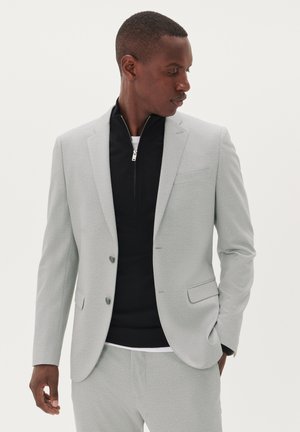 Light gray suit jacket with a textured finish, single-breasted design, two buttons, and flap pockets, paired with a black zip-up top.