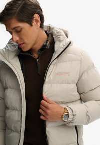 Chaqueta de plumas beige claro con cierre de cremallera y capucha. Suéter marrón texturizado debajo, acompañado de un reloj dorado.