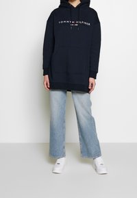 Sudadera oversized en color navy con el logo "TOMMY HILFIGER EST. 1985"; incluye un bolsillo frontal y cordones ajustables. Combinada con vaqueros de pierna ancha en azul claro.