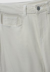 Beżowe spodnie bawełniane o prostym kroju, z białymi szwami, przednią kieszenią i metalowym guzikiem w okolicy talii.