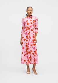 Γυναίκα που φοράει μακρύ ροζ φόρεμα με κουμπιά, με κόκκινα και ροζ κυκλικά σχέδια, και άσπρα σανδάλια με ψηλό τακούνι, στέκεται μπροστά σε απλό φόντο.