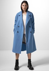 Cappotto blu doppiopetto con ampi rever, tasche laterali e tessuto texturizzato. Indossato sopra un maglione chiaro e una gonna grigia, abbinato a stivali neri.