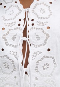 Blusa bianca ricamata con motivi floreali a punto aperto, scollatura rotonda e dettaglio di chiusura con fiocco sul davanti. Tessuto di lino testurizzato.