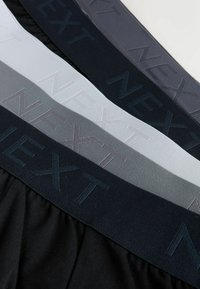 Ensemble de quatre slips à ceinture élastique en noir, gris, blanc et bleu foncé. Chacun comporte l'inscription "NEXT" en relief sur la ceinture. Texture de tissu doux.