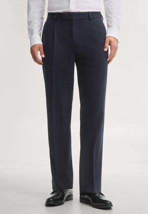 Mężczyzna ubrany w ciemnogranatowe formalne spodnie, czarne eleganckie buty i białą koszulę, stojący na prostym jasnym tle.