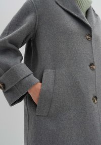 Cappotto di lana grigio con colletto a punta, chiusura frontale con bottoni, tasche laterali e finitura testurizzata.