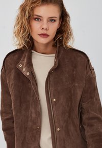 Bruine suède jas met een ritssluiting aan de voorkant, goudkleurige hardware en een ontspannen pasvorm. Opvallende schouderepauletten en een zachte textuur. Gekocht over een beige top.