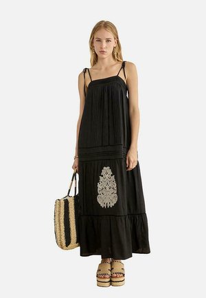 Mujer con vestido negro sin mangas con detalles bordados, sosteniendo un bolso tote tejido a rayas negro y beige, y calzando sandalias de plataforma.