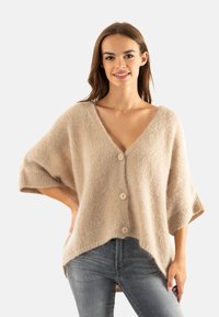 Cardigan en tricot beige avec un col en V, des manches courtes et quatre boutons clairs sur le devant. Tissu doux et texturé. Ourlet asymétrique.