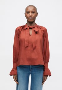 Blusa color ruggine con fiocco al collo, maniche lunghe con polsini arricciati e una vestibilità leggermente oversize.