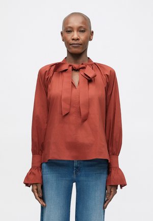 Blouse couleur rouille avec un nœud au cou, manches longues avec des poignets volants et une coupe légèrement ample.