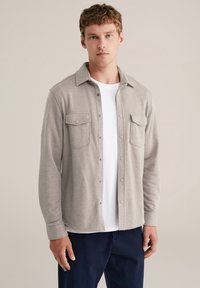 Cămașă deschisă la culoare, dintr-un material moale, cu două buzunare la piept și guler. Purtați peste un tricou alb și pantaloni bleumarin.