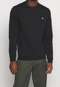Sudadera negra de algodón, con un cuello redondo, puños acanalados y un pequeño logo de flor de lis dorado en el pecho.