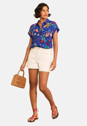 Frau trägt ein blaues T-Shirt mit kurzen Ärmeln und bunten Mustern, weiße Shorts, rote Schnürsandalen und hält eine kleine Korbtasche.