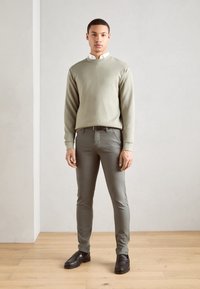 Light green sweatshirt, fitted gray pants, black leather shoes, layered with a white collared shirt, standing on a wooden floor against a gray wall.