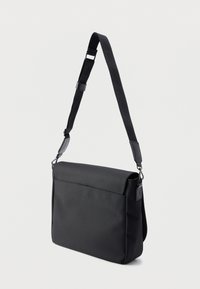 Μαύρο τσαντάκι ταχυμεταφορέα από υφασμάτινη υφή, με αναδιπλούμενο καπάκι, ρυθμιζόμενο ιμάντα και μπροστινή τσέπη.