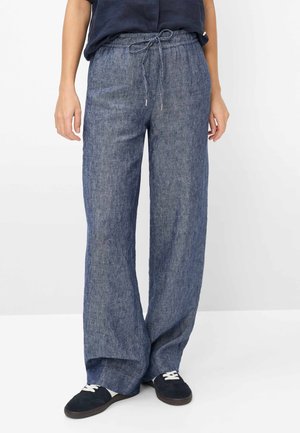 Person trägt lockere blaue Leinenhose mit Kordelzug in der Taille und schwarze Turnschuhe mit weißen Schnürsenkeln, steht vor weißem Hintergrund.