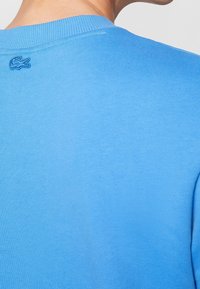 Hombre vistiendo un suéter de cuello redondo de canalé azul claro con un pequeño logo de cocodrilo azul bordado cerca del cuello en la parte trasera.
