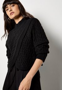 Czarny sweter z dzianiny z teksturowanym wzorem w warkocze, wysoka szyja i skrócone rękawy. Materiał wydaje się miękki i ciepły, idealny do warstwowego ubierania.