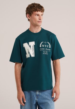 Jeune homme portant un t-shirt oversize turquoise avec un graphisme et texte NYC blancs, associé à un jean bleu clair, regardant sur le côté.