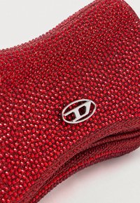 Prim-plan cu o țesătură roșie sclipitoare, texturată cu mici strasuri, și un logo oval argintiu cu litera „D” în centru.