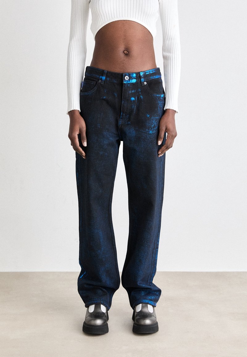 Personne portant un jean large bleu foncé légèrement brillant avec des baskets noires et blanches et un pull court côtelé blanc.