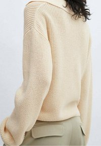 Knit cream sweater with a wide collar and loose fit, featuring ribbed texture and dropped shoulders, paired with khaki pants.