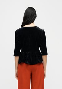 Black velvet peplum top with three-quarter sleeves, featuring a wide neckline and soft texture, paired with red trousers.