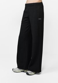 Czarne spodnie o szerokich nogawkach wykonane z miękkiego materiału z elastycznym pasem. Posiadają małe białe logo z boku i są zestawione z jasnoszarymi trampkami.