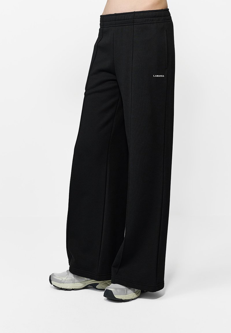 Czarne spodnie o szerokich nogawkach wykonane z miękkiego materiału z elastycznym pasem. Posiadają małe białe logo z boku i są zestawione z jasnoszarymi trampkami.