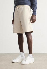 Beige cotton shorts with an elastic waistband and contrasting drawstring. "Calvin Klein" logo in light pink embroidered on the side.