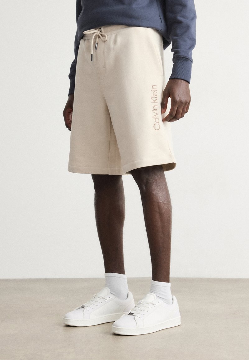 Beige cotton shorts with an elastic waistband and contrasting drawstring. "Calvin Klein" logo in light pink embroidered on the side.