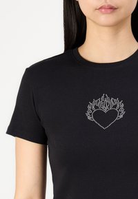 Melnas kokvilnas T-kreklā ir sirds dizains ar liesmu līdzīgiem rotājumiem, kas izgatavoti no rhinestones, uz krūtīm. Klasisks apaļais kakls.