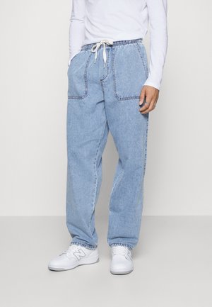 Modelka nosząca jasnoniebieskie joggery dżinsowe z elastycznym pasem i sznurkiem, w połączeniu z białą bluzką z długim rękawem i białymi tenisówkami.